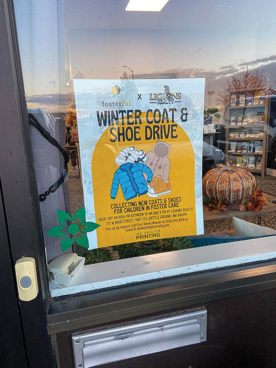 A poster for the winter coat and shoe drive hosted by Fosterful and Legions Realty is displayed in a window.