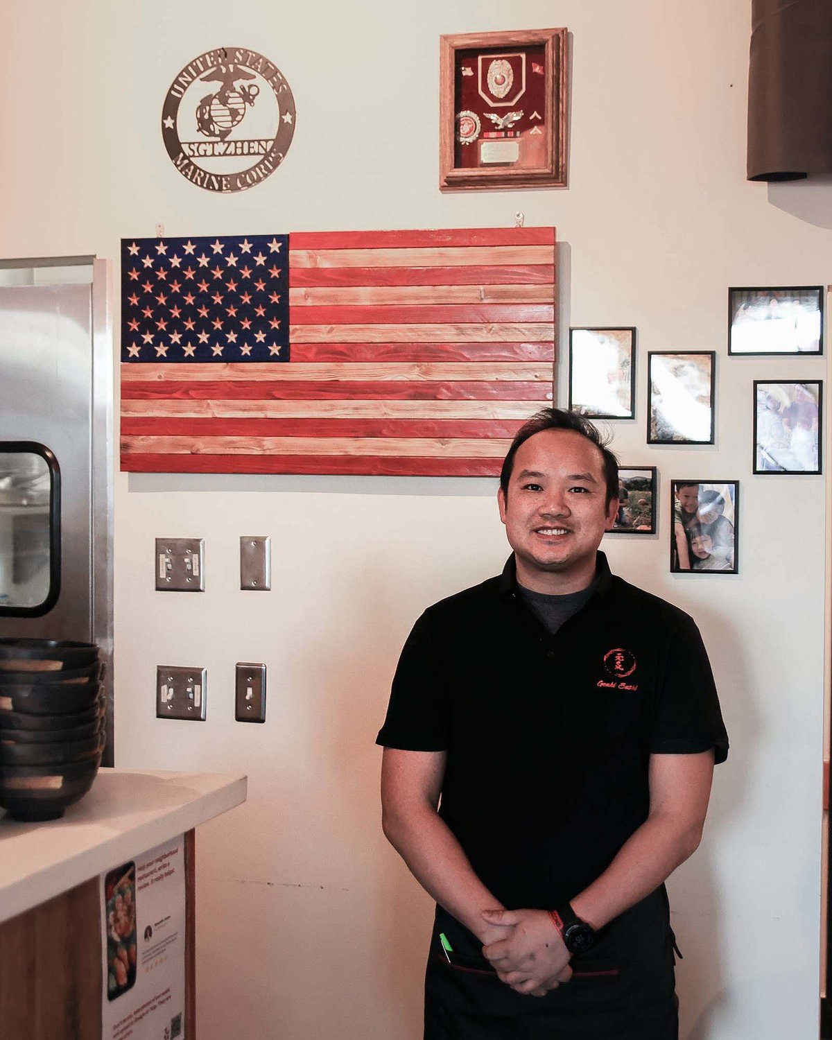 Eddie Zhen, the owner of Genki Sushi in Battle Ground, stands in front of a wall in his restaurant which honors his service to the country and includes photos of his family on Wednesday, Feb. 15.